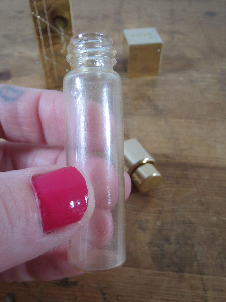 May include: A clear, small glass perfume bottle with a threaded neck. The bottle is held in a hand with red nail polish. A gold-colored perfume bottle cap and a gold-colored box are in the background.