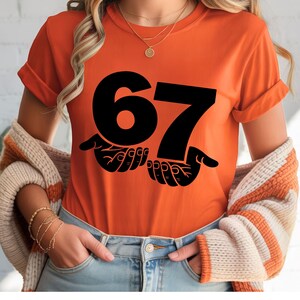 Puede incluir: Camiseta naranja con el n&uacute;mero "67" en negro, sostenida por dos manos negras. Las mangas est&aacute;n dobladas. La persona lleva vaqueros azules y un c&aacute;rdigan color crema.