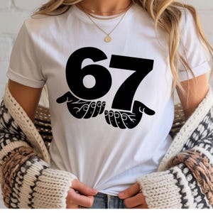 Puede incluir: Camiseta blanca con el n&uacute;mero 67 en negro, sostenido por manos estilizadas. El dise&ntilde;o, centrado en la camiseta, crea un impacto visual sencillo y llamativo.
