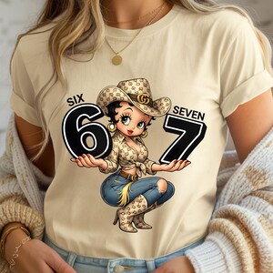 May include: A beige t-shirt featuring a cartoon cowgirl holding the numbers "6" and "7". The cowgirl wears a cowboy hat and a patterned shirt, with blue jeans and boots. The words "SIX" and "SEVEN" are above the numbers.
