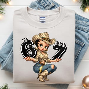 May include: A light beige Gildan sweatshirt featuring a cartoon cowgirl holding the numbers 6 and 7. The cowgirl wears a patterned cowboy hat and shirt, blue jeans, and cowboy boots. The text "SIX" and "SEVEN" are above the numbers.