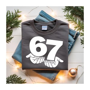 Puede incluir: Una sudadera gris oscuro con el n&uacute;mero 67 en blanco, sostenido por dos manos blancas. La sudadera est&aacute; doblada sobre un par de vaqueros azules. Decoraciones navide&ntilde;as, incluyendo vegetaci&oacute;n y un adorno dorado, rodean la ropa.