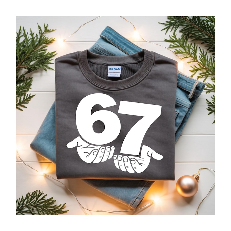 Puede incluir: Una sudadera gris oscuro con el n&uacute;mero 67 en blanco, sostenido por dos manos blancas. La sudadera est&aacute; doblada sobre un par de vaqueros azules. Decoraciones navide&ntilde;as, incluyendo vegetaci&oacute;n y un adorno dorado, rodean la ropa.