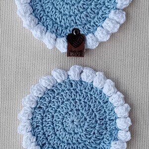 May include: Two circular crochet coasters featuring a light blue center with a textured white puff stitch border. Each coaster includes a small, dark brown heart-shaped wooden tag at the bottom edge with the text Made with LOVE engraved.