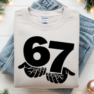 Puede incluir: Una sudadera color crema con el n&uacute;mero "67" en negrita negra. El n&uacute;mero est&aacute; sostenido por dos manos negras. La sudadera est&aacute; doblada sobre un par de vaqueros azules.