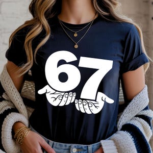 Puede incluir: Una camiseta azul marino con el n&uacute;mero "67" en letras blancas grandes, sostenida por dos manos blancas. La camiseta tiene mangas cortas y cuello redondo.