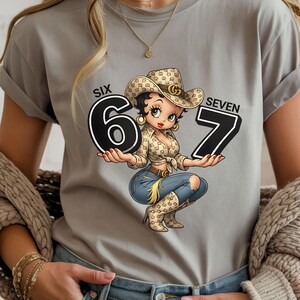 May include: A gray t-shirt featuring a cartoon cowgirl holding the numbers 6 and 7. The cowgirl wears a cowboy hat, a patterned shirt, and blue jeans. The words "SIX" and "SEVEN" are above the numbers.