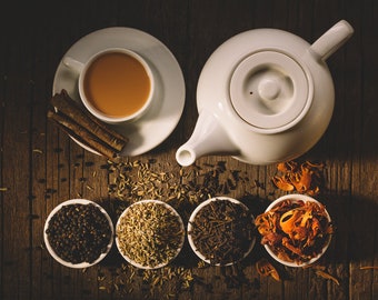 May include: A white teapot with a lid and a cup of tea on a wooden surface with various spices, including cinnamon sticks, black peppercorns, fennel seeds, and nutmeg.