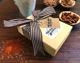 May include: A gift box wrapped in brown and white striped ribbon with a brown tag that reads "The Tea Crew Premium Loose Leaf Tea" on a wooden surface with a cup of coffee and bowls of nuts.