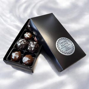 Puede incluir: Una caja negra con una etiqueta plateada que dice "Cocoa Elite Pure Edible Silver Leaf Luxury Chocolates" está abierta, revelando una caja de ocho trufas de chocolate con acentos de hoja plateada.