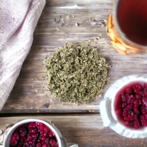 May include: Overhead view of a wooden surface featuring a pile of dried green herbs, a glass of red liquid, and two small bowls filled with red berries. A white cloth is partially visible on the left.