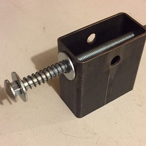 May include: A metal spring-loaded mechanism with a threaded rod and a round knob. The mechanism is attached to a rectangular metal box with two holes.