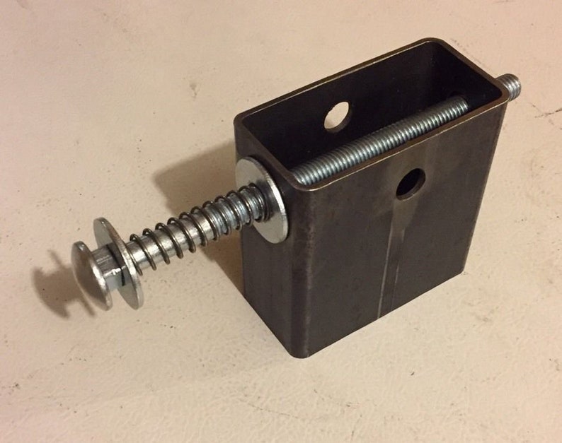 May include: A metal spring-loaded mechanism with a threaded rod and a round knob. The mechanism is attached to a rectangular metal box with two holes.