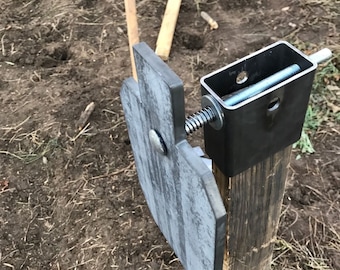 May include: A metal shooting target attached to a wooden post with a metal bracket and a screw. The target is grey and has a rectangular shape with a rounded top.