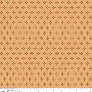 May include: A light orange fabric featuring a repeating pattern of small, dark brown four-petaled floral shapes. A white ruler is visible at the bottom edge, showing a scale of 12 inches, with the text Riley Blake Designs printed on it.