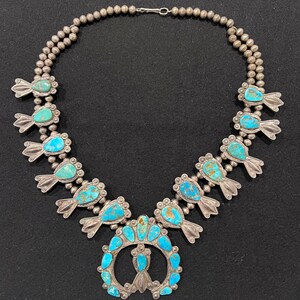 May include: A silver and turquoise necklace with a large, ornate pendant. The pendant is shaped like a horseshoe and is decorated with turquoise stones. The necklace is made of silver beads and has a clasp.