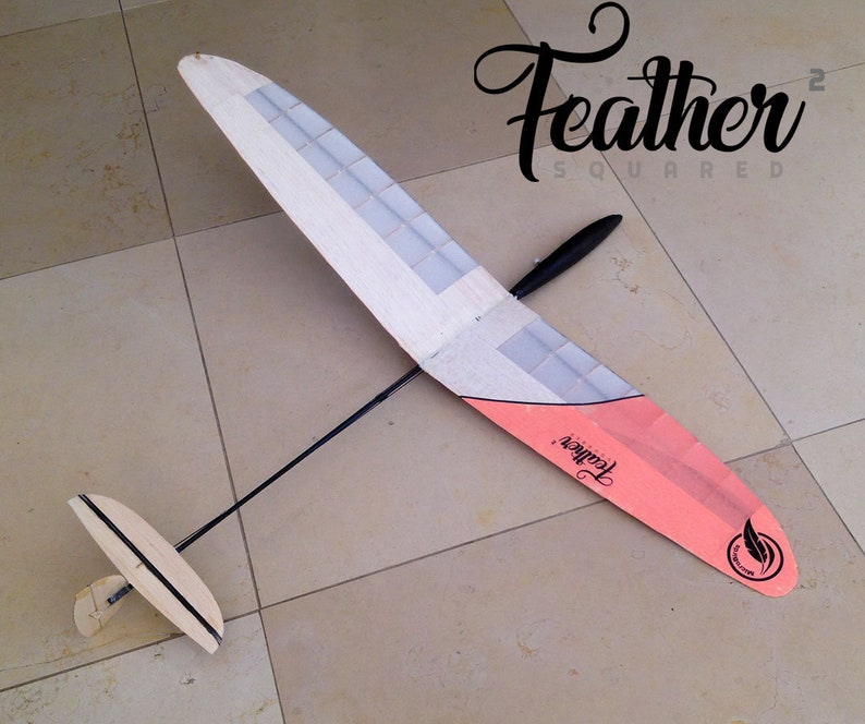 Puede incluir: Un modelo de planeador con un ala de madera clara y una punta de ala rosa y blanca. El texto "Feather Squared" est&aacute; en escritura negra. El planeador tiene una cola negra y un ala peque&ntilde;a en la parte trasera.