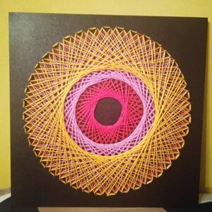 May include: A circular string art piece with a concentric design. The outer ring is yellow, the middle ring is pink, and the inner ring is red. The piece is mounted on a black background.