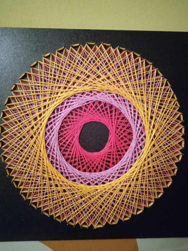May include: A circular string art piece with a yellow, pink and red design. The string is wrapped around nails hammered into a black surface.