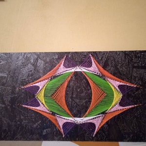 May include: A colorful string art piece with a geometric design. The design is made with different colored strings, including purple, pink, orange, green, and yellow. The strings are stretched across a black wooden board.