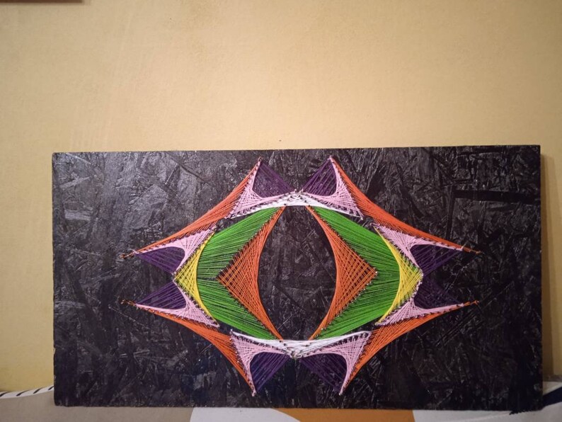 May include: A colorful string art piece with a geometric design. The design is made with different colored strings, including purple, pink, orange, green, and yellow. The strings are stretched across a black wooden board.