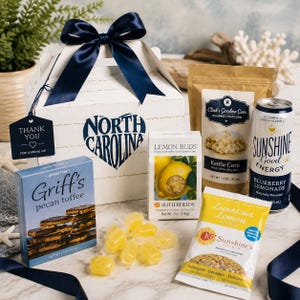 May include: A white gift box with a dark blue ribbon and a North Carolina logo, containing Griff's pecan toffee, Butterfields lemon buds, Clark's Carolina Corn kettle corn, a Sunshine energy drink, and a KC Sunshine's lemon cookie with loose yellow candies.