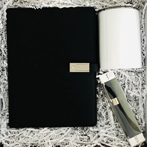 May include: A black notebook with a silver clasp, a white tumbler, and a pen in a clear case are arranged in a gift box filled with white paper shreds. The notebook has a debossed design. The pen case has text.