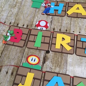 May include: A brown and green banner with the words "Happy Birthday Jax" in colourful letters. The banner is decorated with Mario characters and objects from the Super Mario Bros. video game.