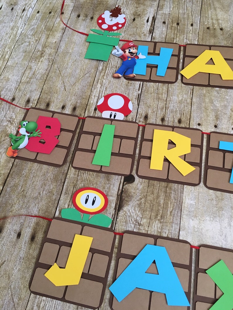 May include: A brown and green banner with the words "Happy Birthday Jax" in colourful letters. The banner is decorated with Mario characters and objects from the Super Mario Bros. video game.