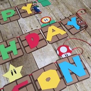 May include: A birthday banner featuring the words "Happy Birthday Son" in colourful letters. The banner is decorated with characters and objects from the Super Mario Bros. video game, including Mario, a Goomba, a Piranha Plant, a mushroom, and a star.