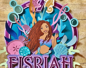 May include: Purple and blue mermaid themed cake topper with a mermaid, a number 7, and the name "EISRIAH".