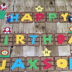 May include: A colourful banner with the words "Happy Birthday Jackson" in a Mario theme. The banner is made of brown cardboard with red, yellow, green, and blue letters. There are also images of Mario characters and items from the game, such as mushrooms, stars, and pipes.