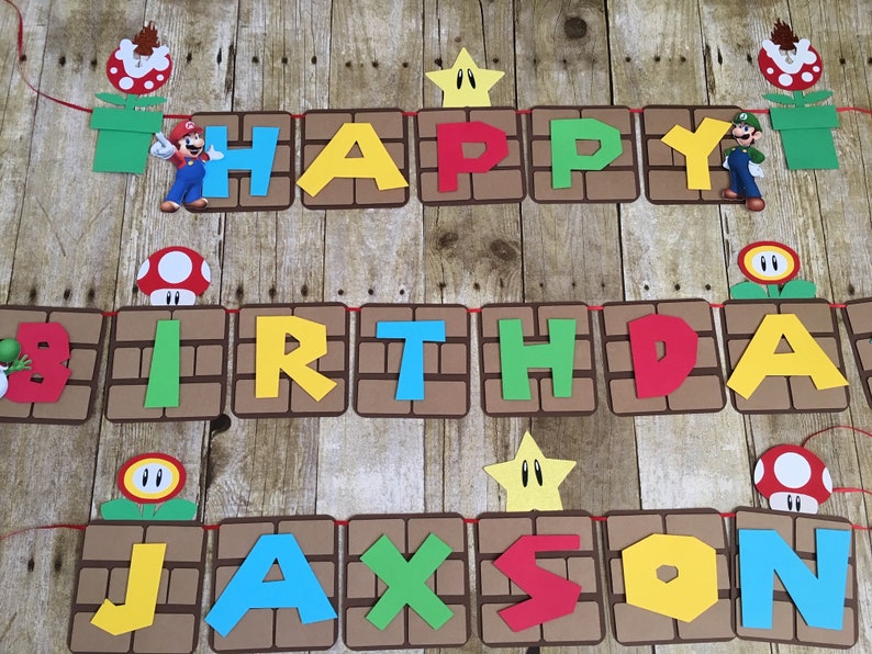 May include: A colourful banner with the words "Happy Birthday Jackson" in a Mario theme. The banner is made of brown cardboard with red, yellow, green, and blue letters. There are also images of Mario characters and items from the game, such as mushrooms, stars, and pipes.