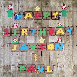 May include: A colourful banner with the words "Happy Birthday Jaxson & Paul" in a Mario-themed design. The banner is made of brown cardboard with red, yellow, green, and blue letters. There are also small Mario characters and objects on the banner, such as a star, a mushroom, and a pipe.