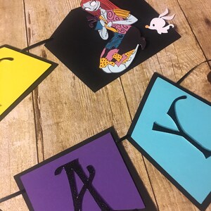 Op de afbeelding: Een zwarte, paarse, blauwe en gele papieren banner met het woord "Happy" in zwarte letters. De banner heeft een uitsnede van Sally uit The Nightmare Before Christmas die een zwarte kat vasthoudt.