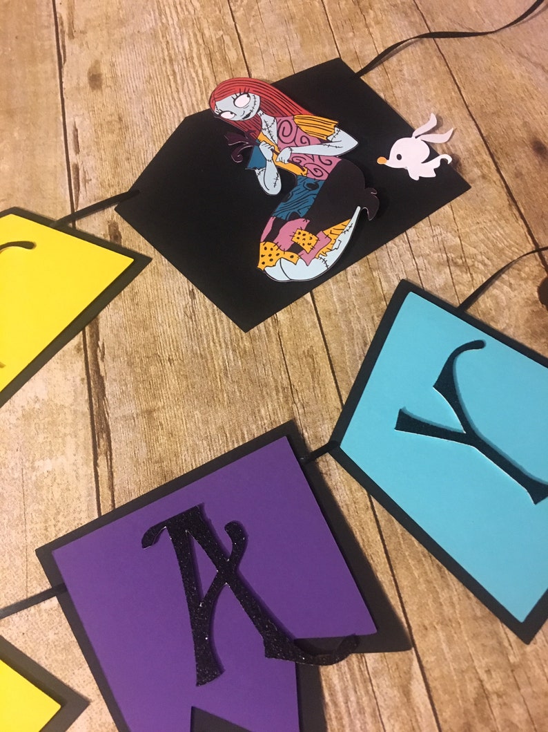 Op de afbeelding: Een zwarte, paarse, blauwe en gele papieren banner met het woord "Happy" in zwarte letters. De banner heeft een uitsnede van Sally uit The Nightmare Before Christmas die een zwarte kat vasthoudt.