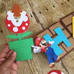 May include: A colourful paper banner featuring Mario, a green pipe, a red mushroom, and the letters "Happy" in blue and brown. The banner is made of cardboard and is perfect for a birthday party.