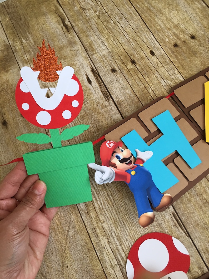 May include: A colourful paper banner featuring Mario, a green pipe, a red mushroom, and the letters "Happy" in blue and brown. The banner is made of cardboard and is perfect for a birthday party.