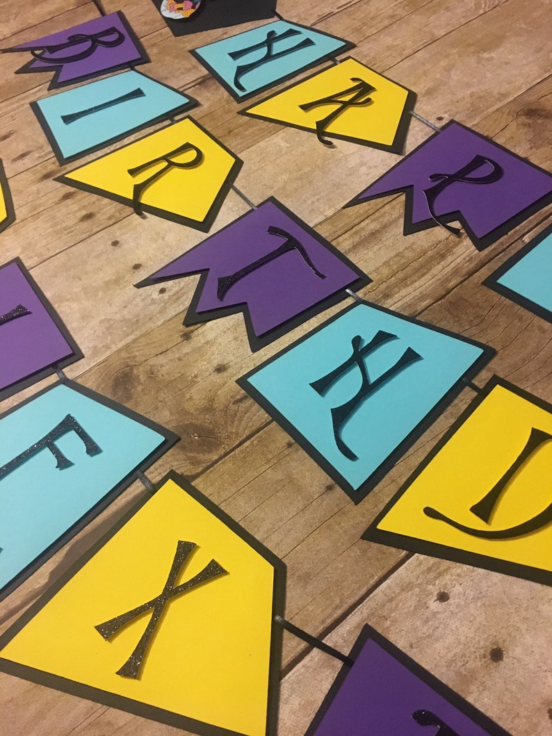 Op de afbeelding: Een kleurrijke verjaardagsbanners met het woord "Birthday" gespeld in zwarte letters op gele, paarse en blauwe vlaggetjes. De vlaggetjes zijn aan elkaar geregen met zwart touw.