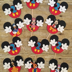 May include: Twelve cupcake toppers featuring Mickey Mouse characters. Each topper has a red, black, and yellow Mickey Mouse head with white gloves. The characters include Mickey Mouse, Minnie Mouse, Donald Duck, Goofy, Pluto, and Daisy Duck. The toppers are decorated with blue glitter numbers "2".