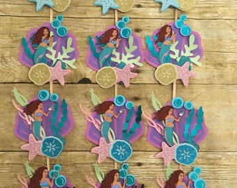 May include: Twelve purple, green, and blue mermaid-themed cupcake toppers with a number six, a mermaid, and seashells.