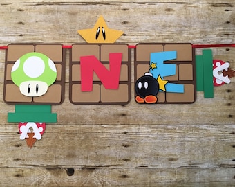 May include: A brown, green, red, blue, and yellow paper banner with the word "ONE" in red letters. The banner is decorated with a mushroom, a star, a bomb, and a pipe. The banner is perfect for a first birthday party.