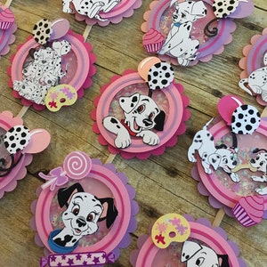 May include: Set of 10 Dalmatian cupcake toppers featuring various images of Dalmatian puppies, including a Dalmatian puppy holding a balloon, a Dalmatian puppy with a lollipop, and a Dalmatian puppy with a paint palette. The toppers are pink and purple with glitter accents.