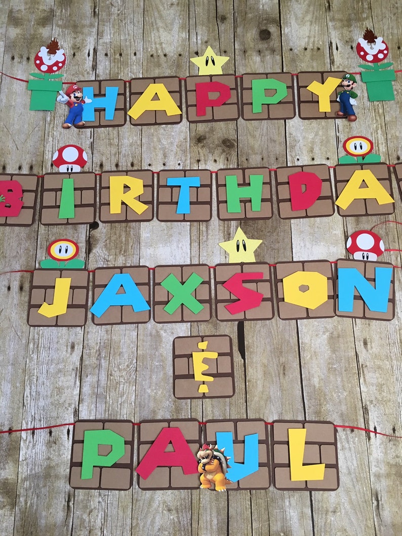 May include: A brown and red banner with the words "Happy Birthday" and a name, "Jaxson Paul", in colourful letters. The banner is decorated with Mario characters and other video game-themed elements.