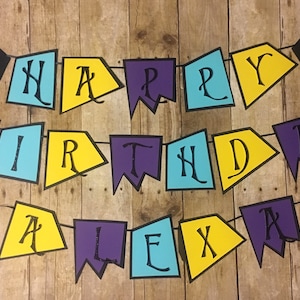 Op de afbeelding: Een kleurrijk spandoek met de woorden "Happy Birthday Alex" in zwarte letters op gele, blauwe en paarse driehoekige vlaggen. Het spandoek is met zwart garen geregen en hangt aan een houten achtergrond.