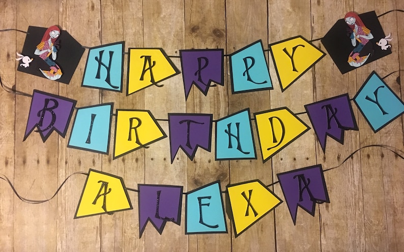 Op de afbeelding: Een kleurrijk spandoek met de woorden "Happy Birthday Alex" in zwarte letters op gele, blauwe en paarse driehoekige vlaggen. Het spandoek is met zwart garen geregen en hangt aan een houten achtergrond.