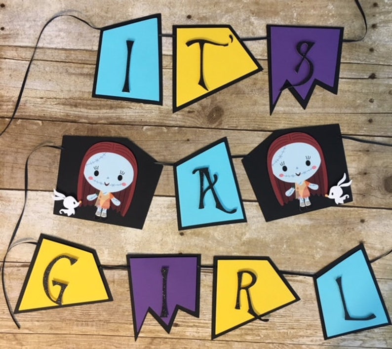 Op de afbeelding: Een 'Het is een meisje'-banner met zwarte, gele, blauwe en paarse vlaggetjes. Elk vlaggetje heeft een letter of een personage uit The Nightmare Before Christmas. De personages zijn Sally en Jack Skellington.