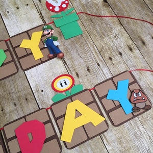 May include: A brown and tan paper banner with the word "Happy" spelled out in red, yellow, and blue letters. The banner is decorated with characters from the video game Super Mario Bros., including a green and blue Mario figure, a red and white flower, and a brown and orange Goomba figure.