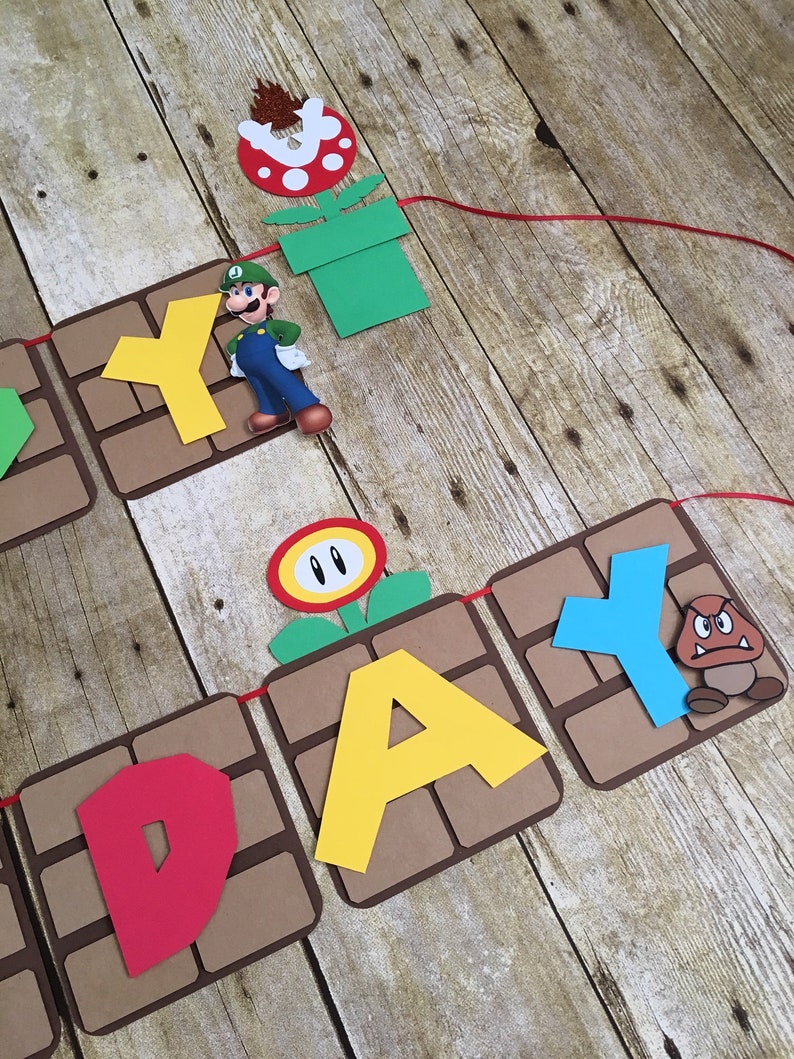 May include: A brown and tan paper banner with the word "Happy" spelled out in red, yellow, and blue letters. The banner is decorated with characters from the video game Super Mario Bros., including a green and blue Mario figure, a red and white flower, and a brown and orange Goomba figure.