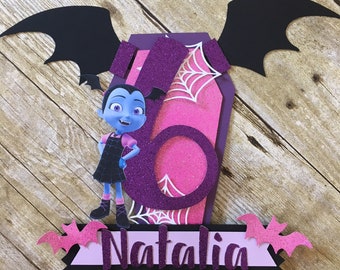 Puede incluir: Un adorno para pastel morado y rosa con una niña vampiro de dibujos animados con alas negras, una telaraña rosa y el nombre "Natalia".
